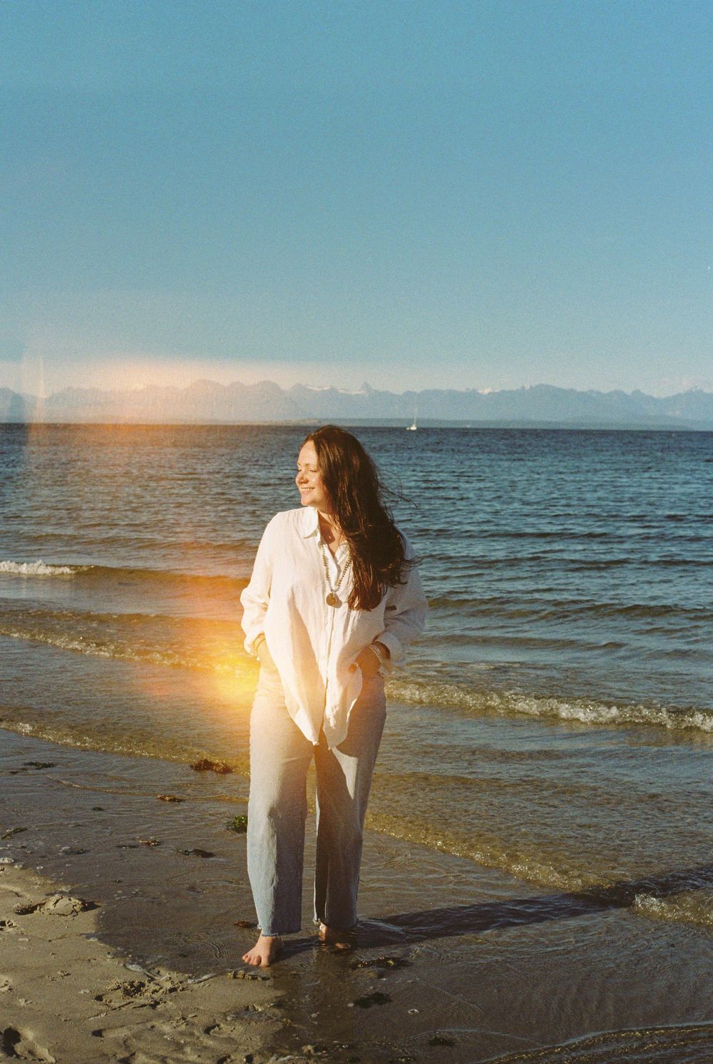Young woman in white blouse and jeans standing on sunny beach shoreline with mountains in background.