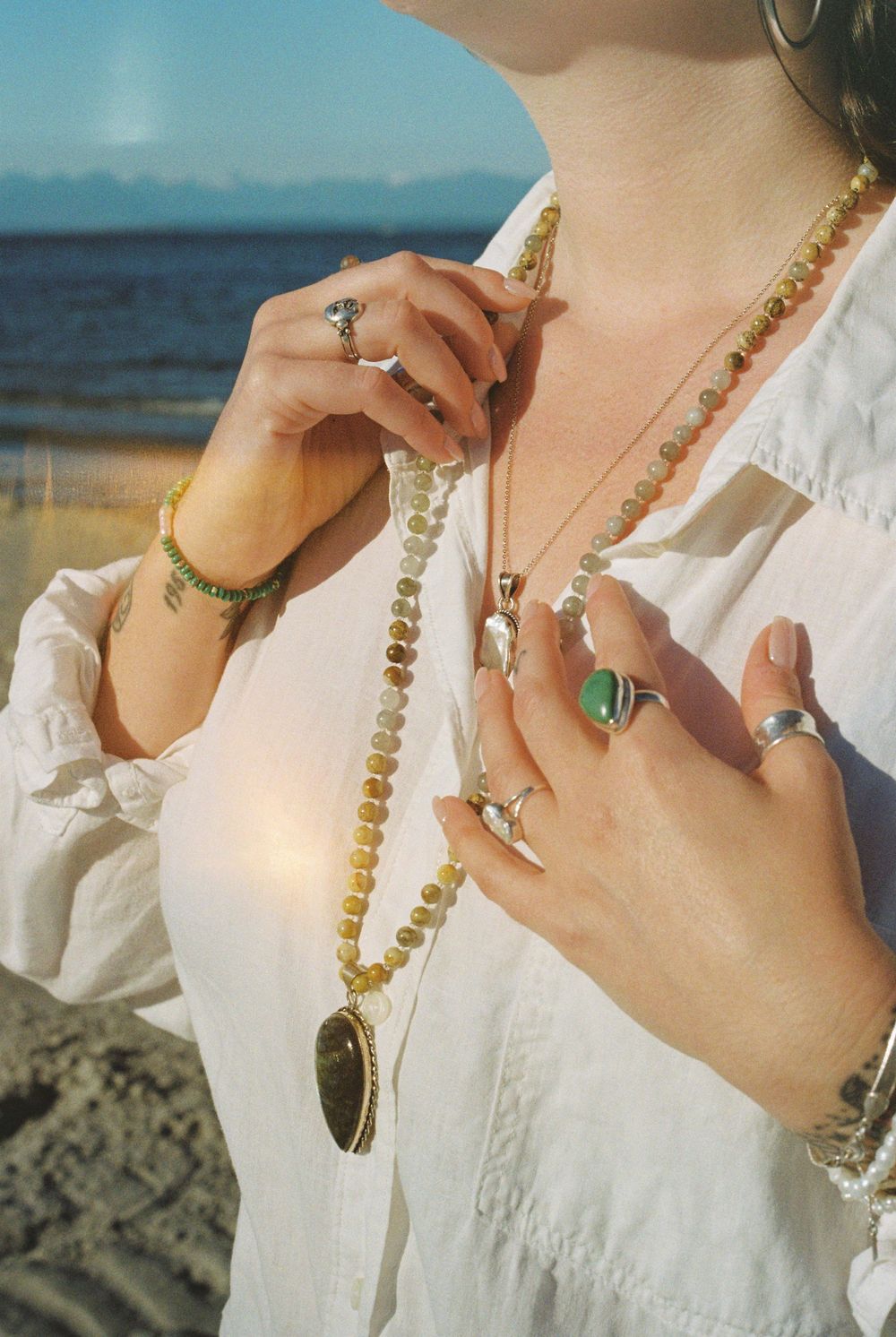 Woman wearing layered gold and green gemstone jewelry, beaded necklace, rings, and bracelets at a beach.