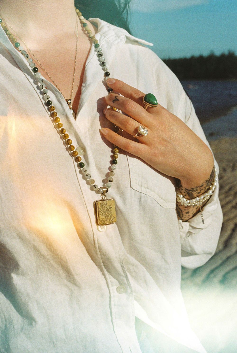 Close-up of layered stone bead necklace with gold pendant and green gemstone ring on woman outdoors.