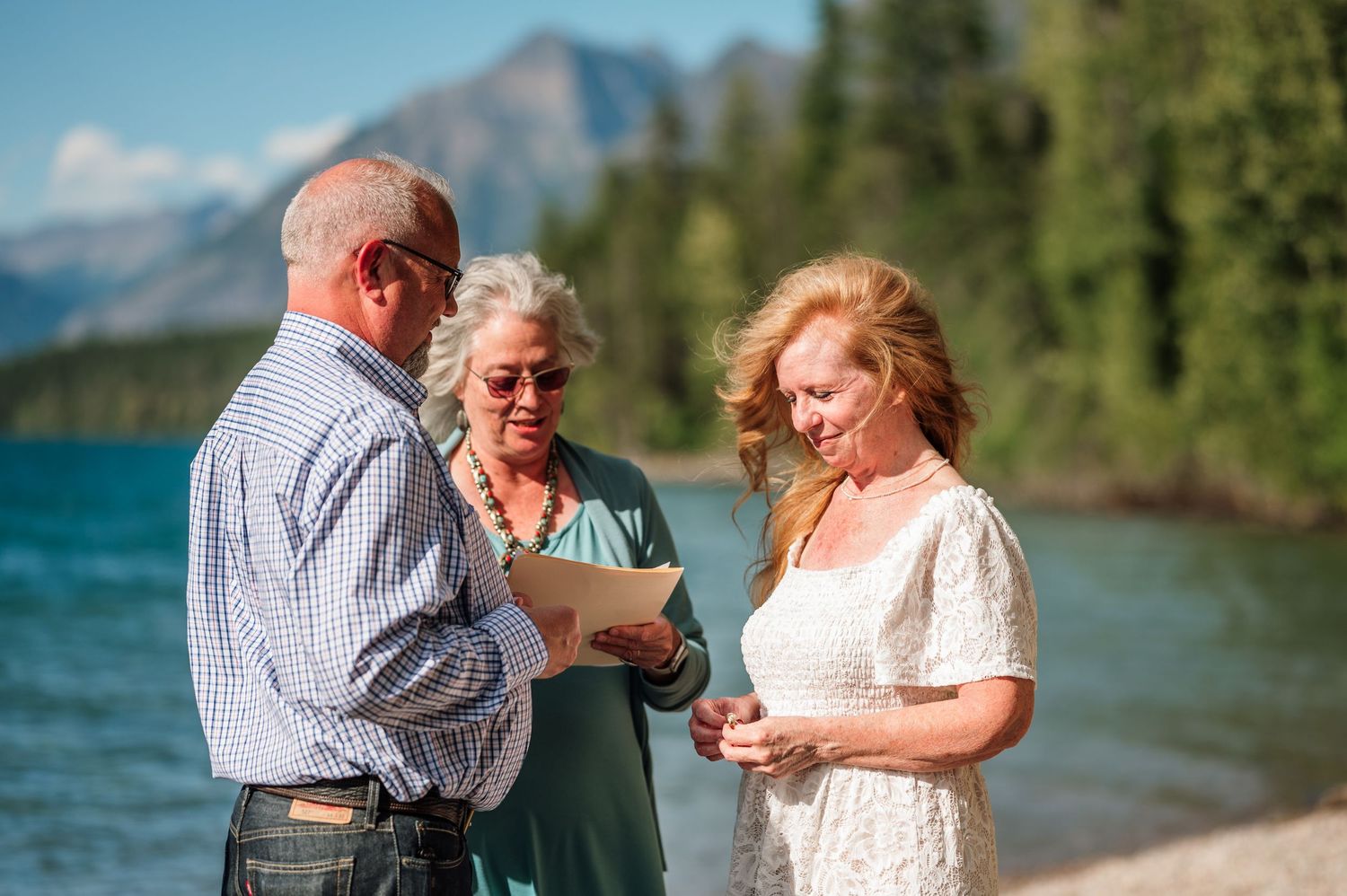Outdoor lakeside wedding ceremony with couple exchanging vows, officiant reading, mountains and trees in background.