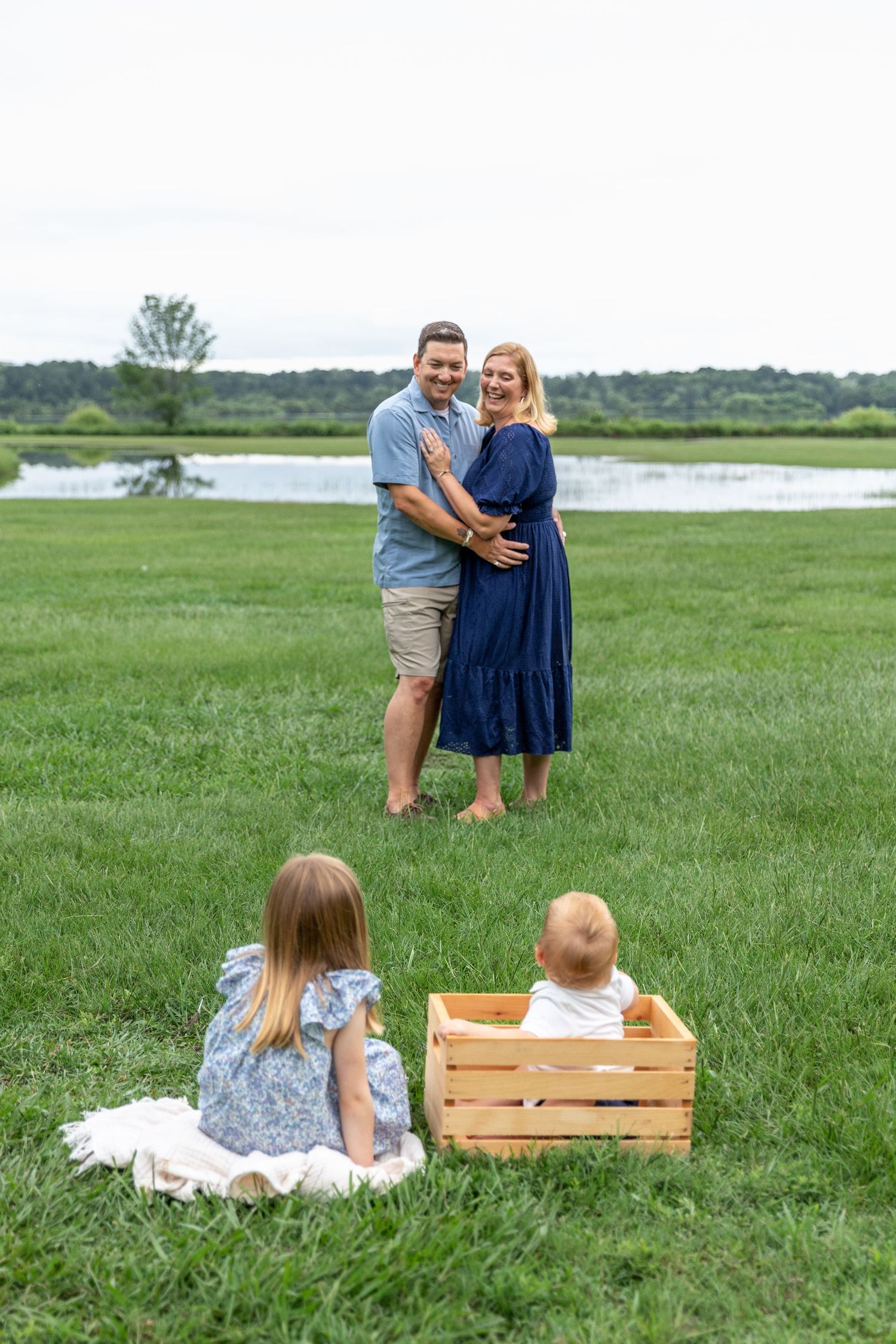 A happy couple embraces on green grass near a lake while two young children sit in the foreground during a family photo session.