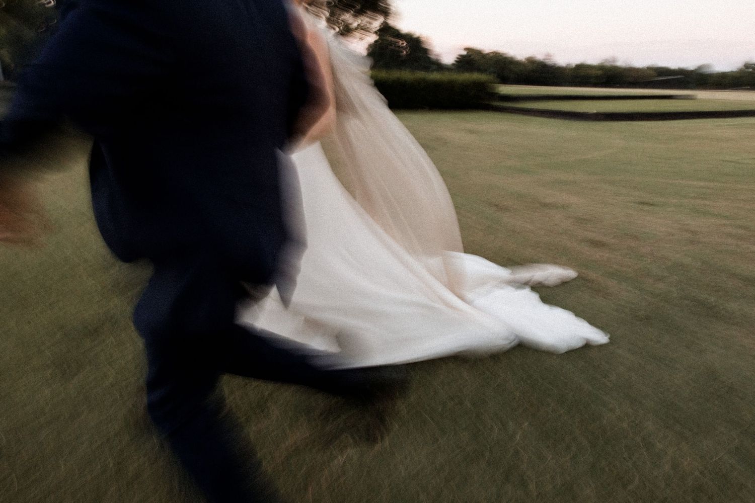 Blurred motion shot of bride and groom running across green lawn at dusk in wedding attire.