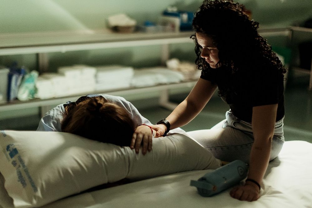 A woman leans over a hospital bed comforting a patient lying on a pillow in a dimly lit medical room.