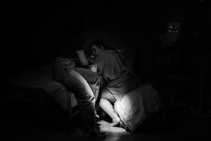 Black and white photo of two people sitting closely together on a bed in a dimly lit room.