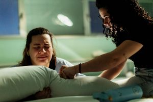 A distressed woman clutches a pillow in a hospital bed while another woman offers comfort and support.