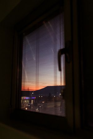 View through a dark window with blinds revealing a colorful sunset sky over a city skyline at dusk.