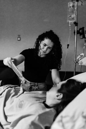 Black and white photo of a woman with curly hair holding hands with a patient lying in a hospital bed with an IV drip.