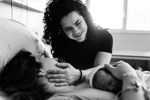 Black and white photo of a smiling woman tenderly touching a new mother holding her newborn baby in a hospital bed.