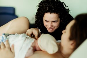 A curly-haired woman smiles lovingly at her newborn baby while resting in a hospital bed after childbirth.
