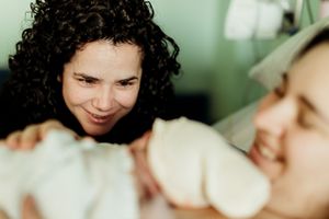 Two women smiling and bonding over a newborn baby wrapped in white blankets in a hospital room.