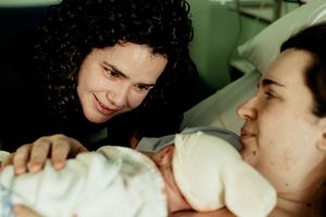 A woman with curly hair smiles at a newborn baby held by a mother lying in a hospital bed after childbirth.