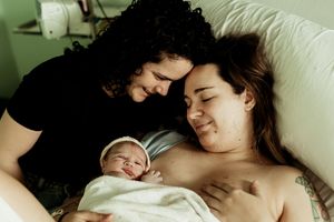 New parents smiling tenderly over their newborn baby wrapped in white blanket in hospital room.