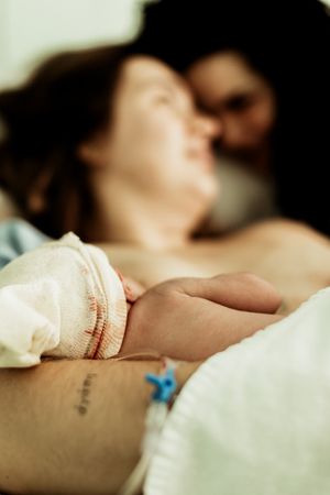 Newborn baby in hospital bracelet resting on mother's arm after birth, with parents sharing tender moment in background.