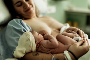 A smiling mother holds her newborn baby on her chest during skin-to-skin contact after birth in a hospital.