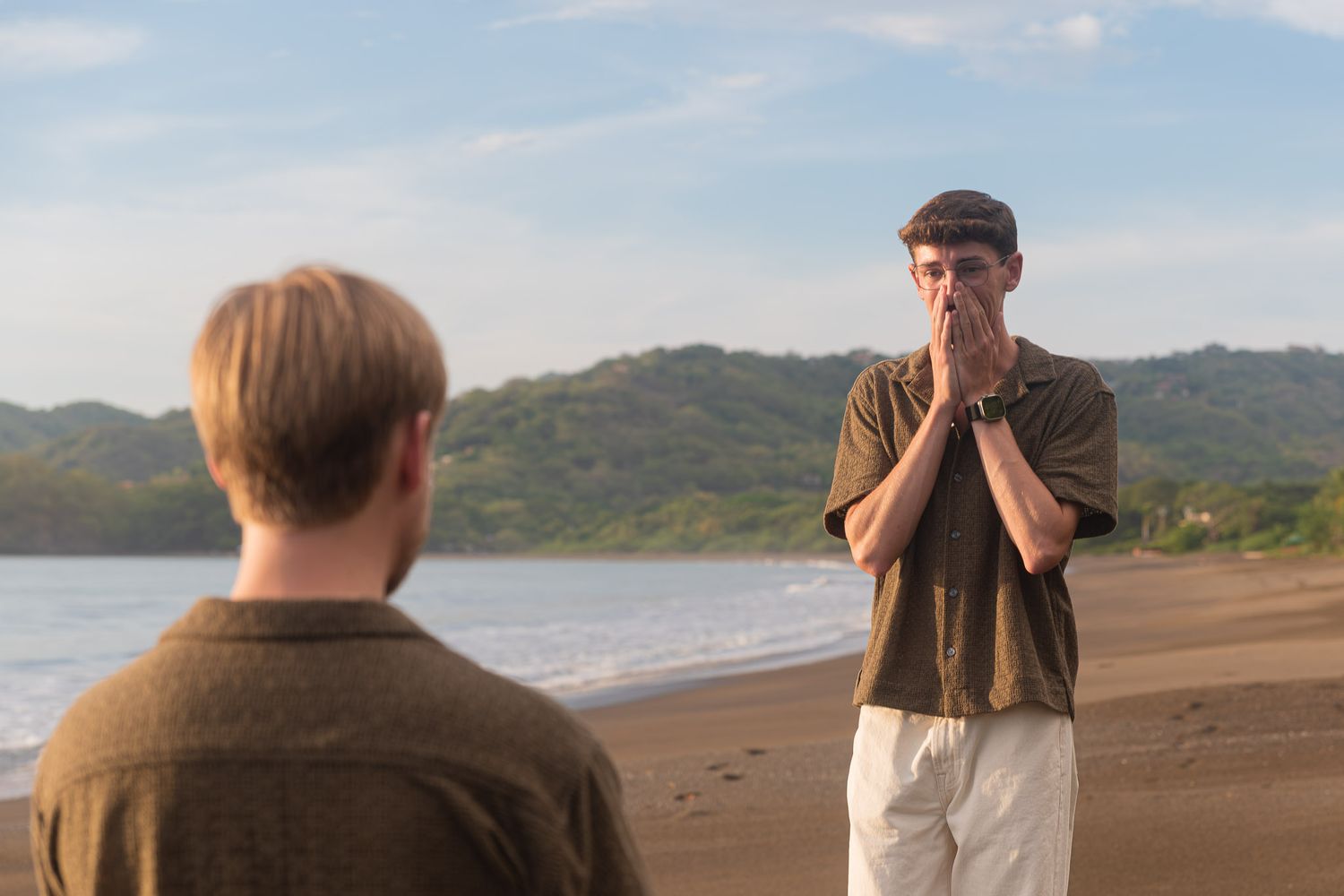 Two men on a tropical beach, one with hands clasped, lush green hills and ocean in the background.