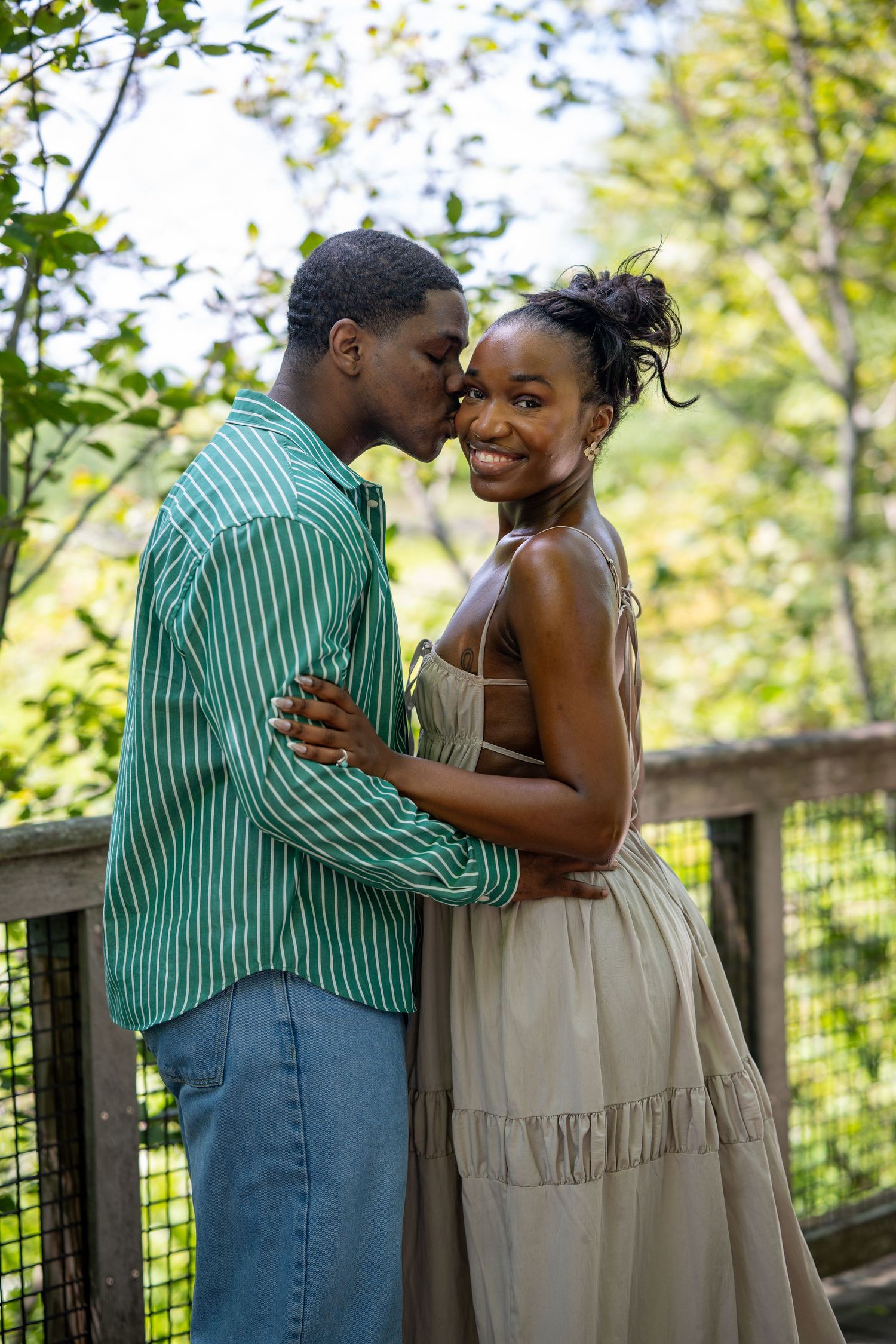 A couple shares a tender kiss on a wooden bridge surrounded by lush green trees in an outdoor park setting.