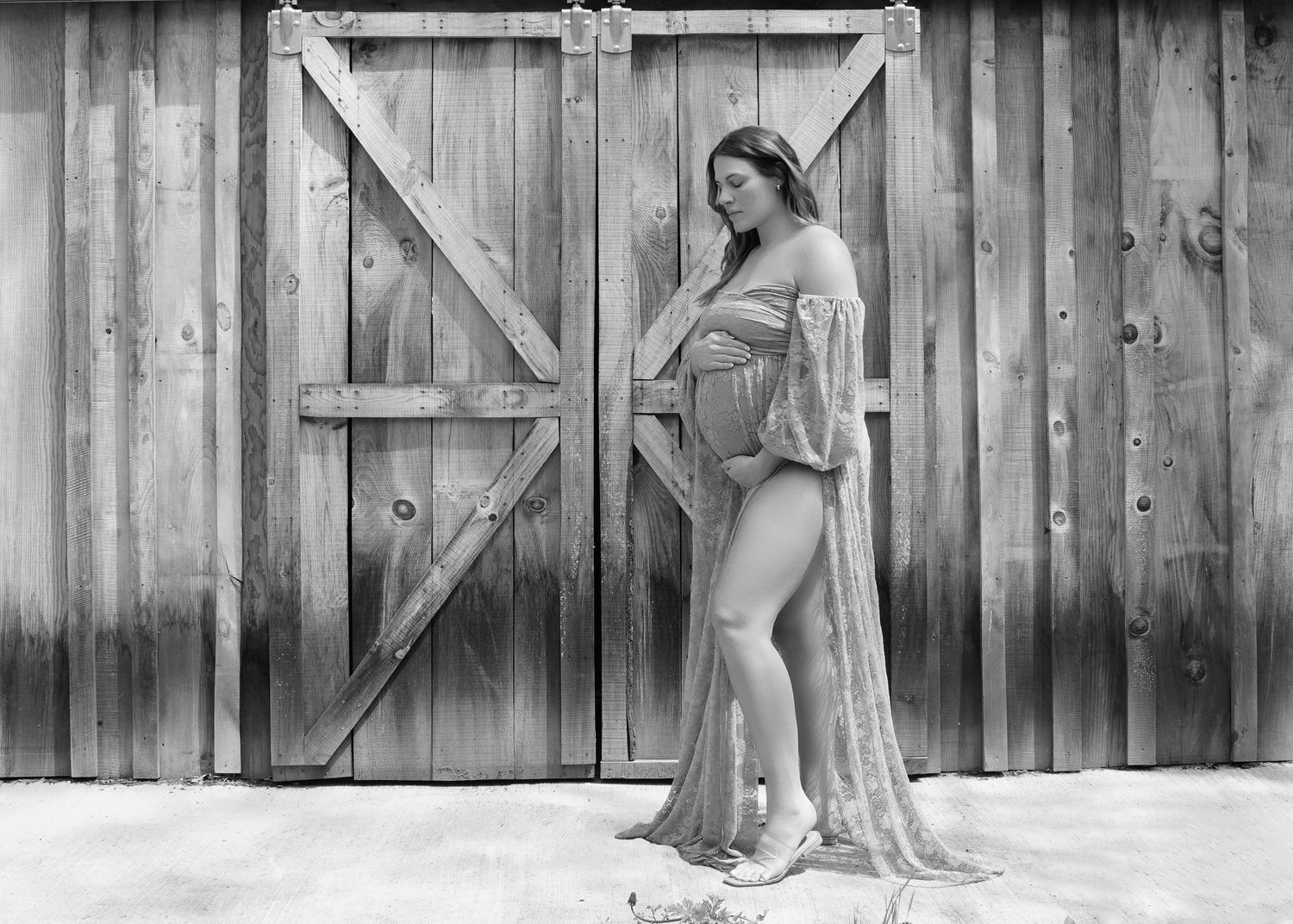 Black and white maternity photo of a pregnant woman in a sheer robe posing in front of rustic barn doors.