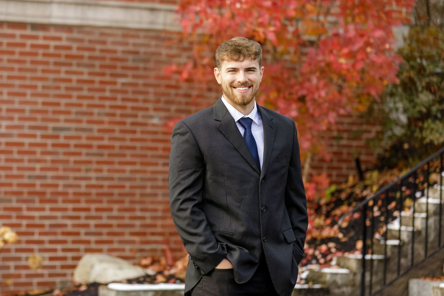 Young professional man in dark suit smiling outdoors against brick wall with autumn red foliage background.