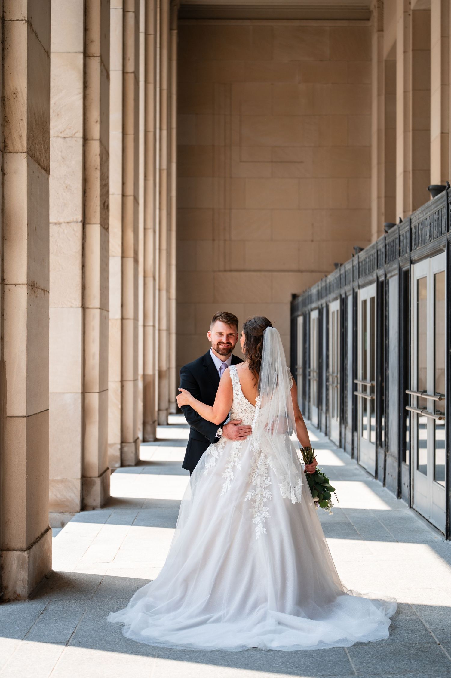 Bride and groom embrace among grand stone columns, her lace gown trailing elegantly as they share a romantic wedding portrait.