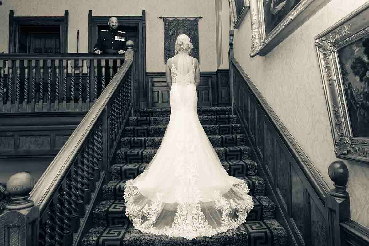 Bride in elegant lace mermaid wedding gown stands on grand staircase with groom watching from above, black and white photo.