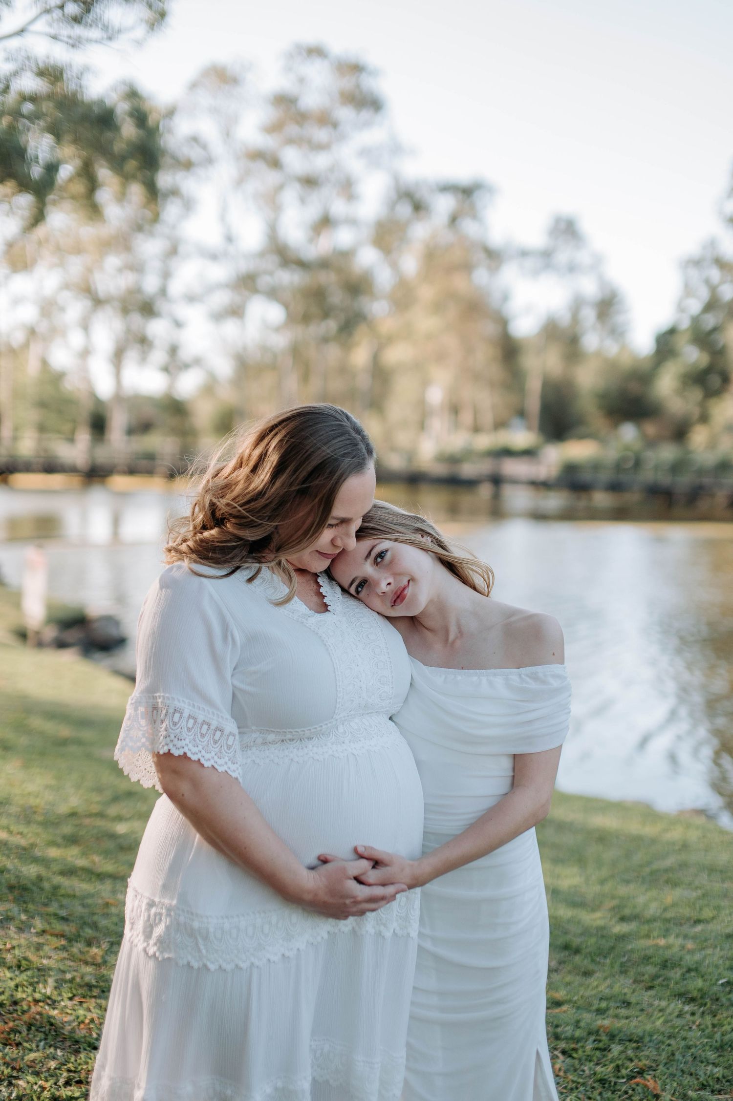 Two women in white dresses share a tender maternity moment by a serene lake surrounded by lush green trees.