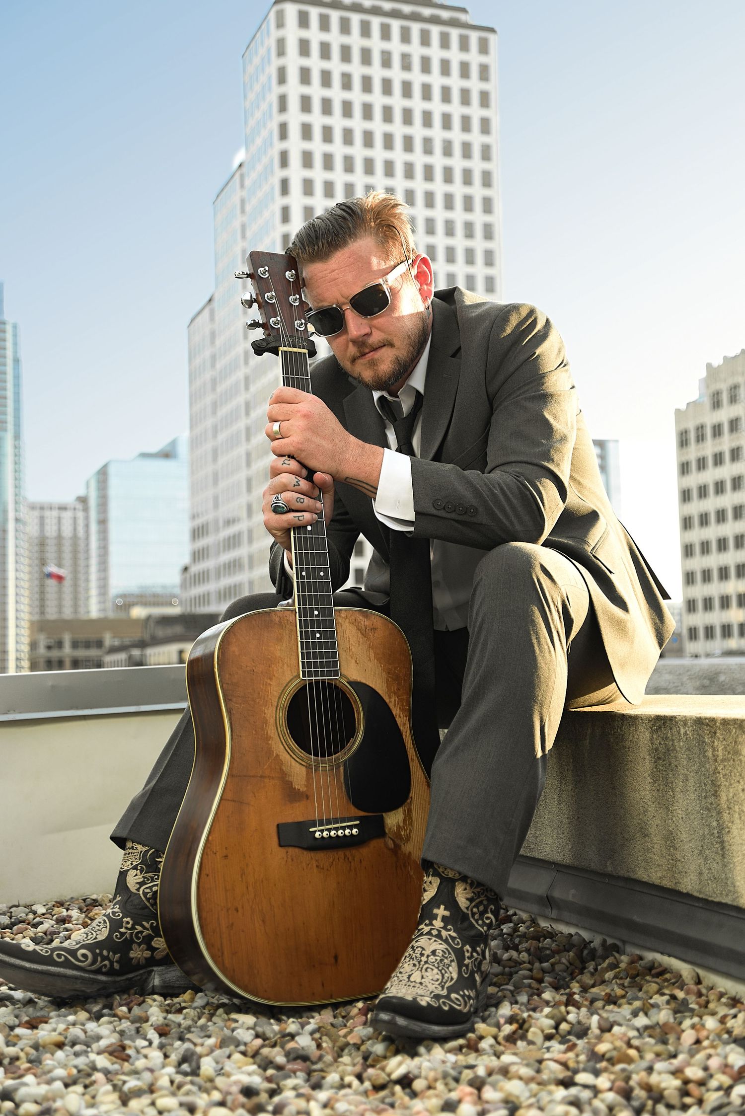 Musician in suit and sunglasses sits on rooftop ledge with acoustic guitar, city skyline behind him.