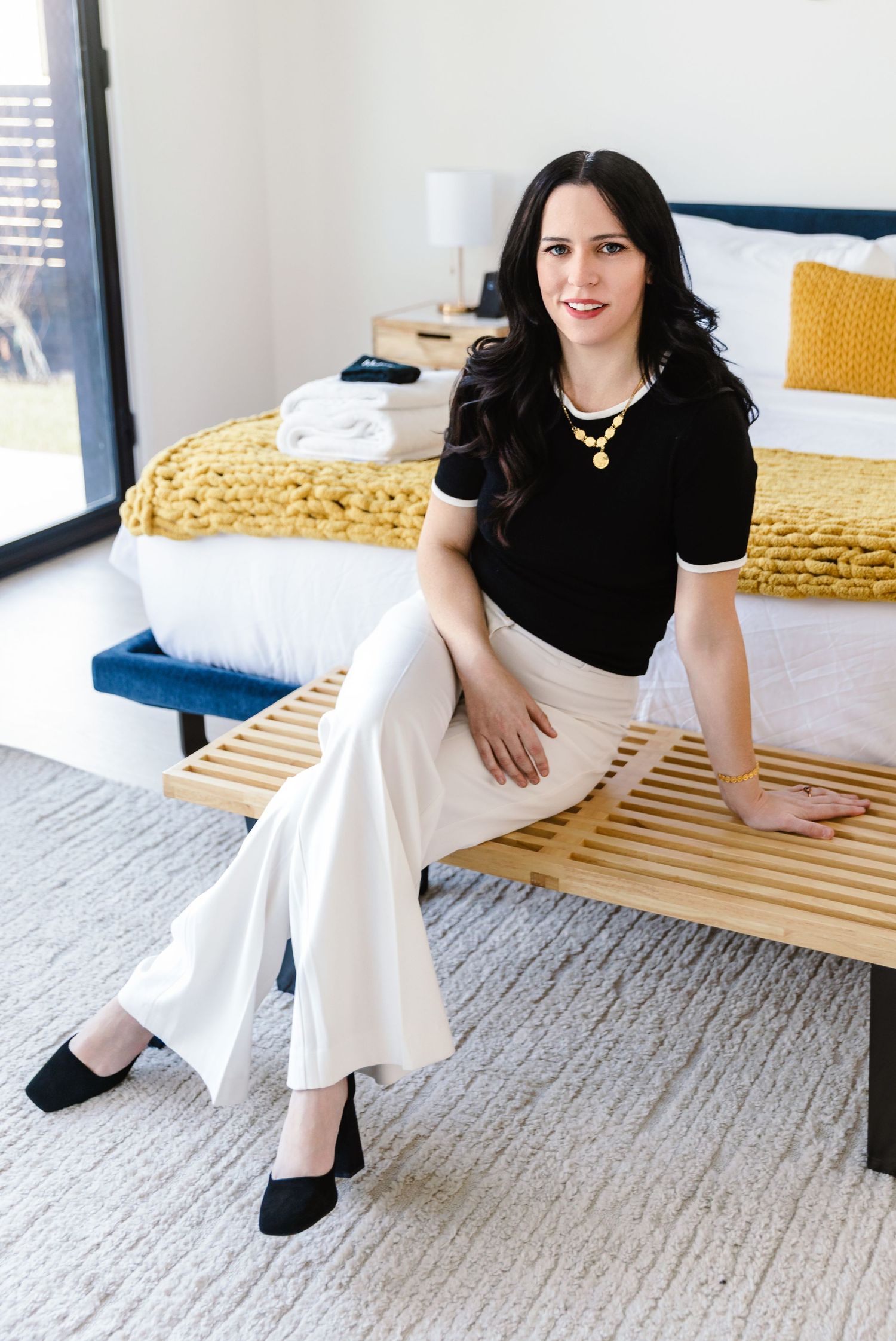 Woman in black top and white wide-leg pants seated on wooden bench in modern bedroom with yellow accents.
