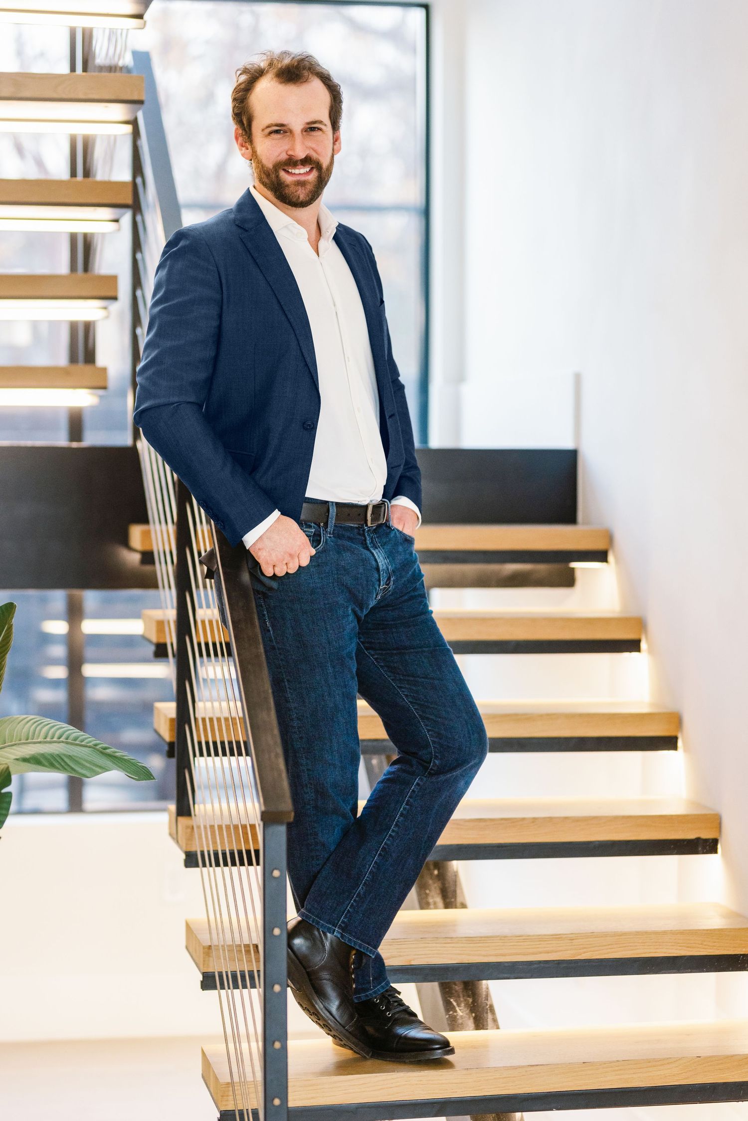 Smiling bearded man in navy blazer and jeans leaning on modern staircase railing in bright office.