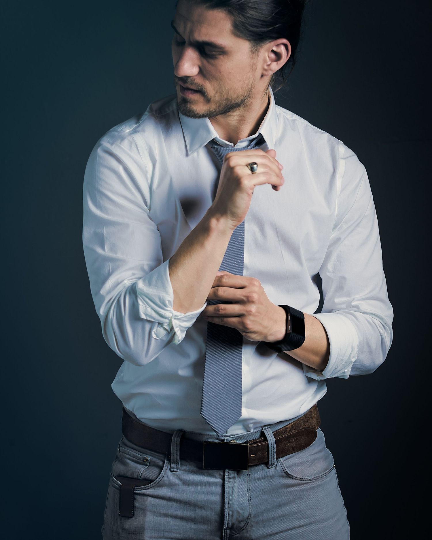 Man in white dress shirt adjusting tie, wearing jeans and black watch, posed against dark background.