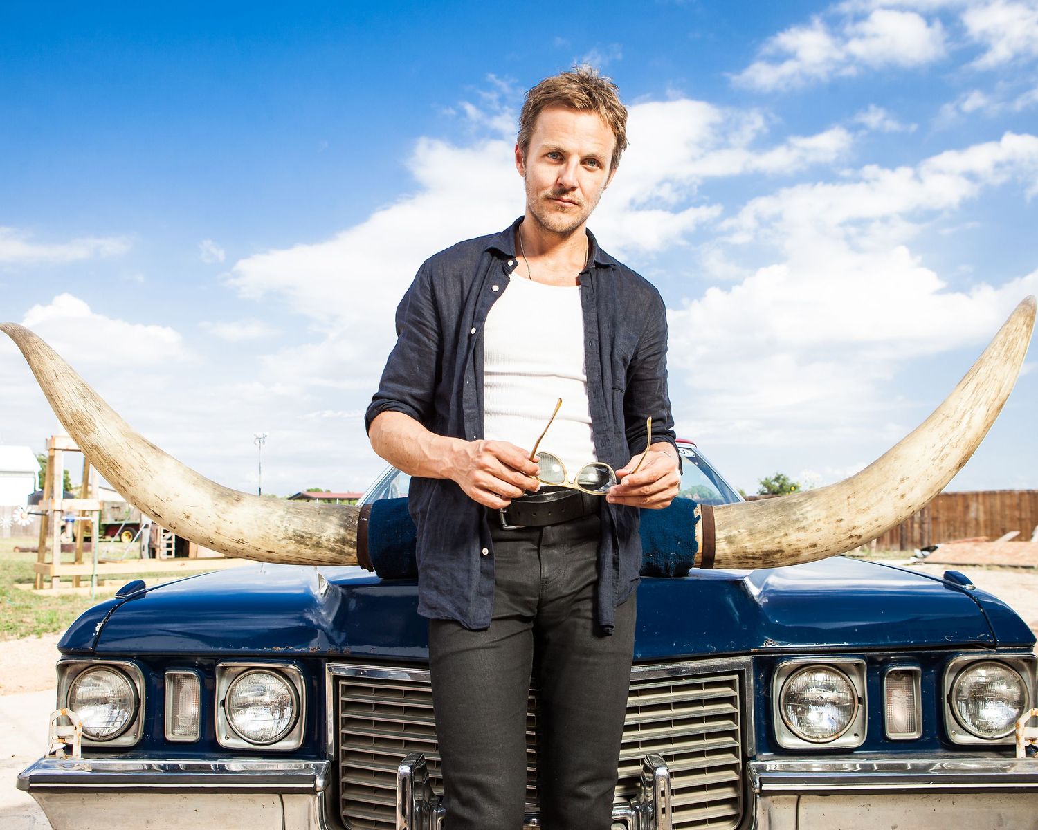 Man leaning on vintage blue car with large longhorn decorations under a blue sky.
