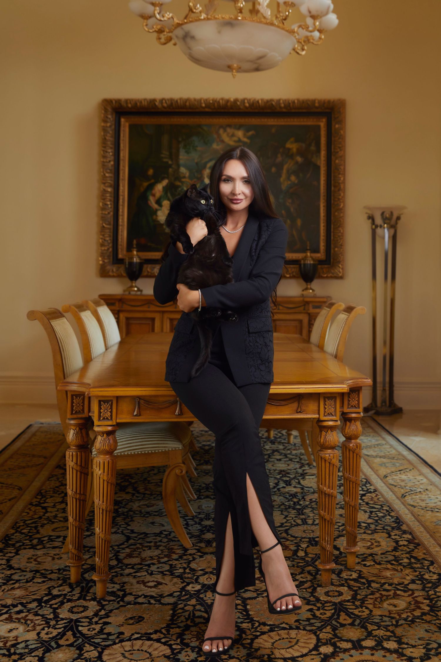 Elegant woman in black suit holding a dark cat in a luxurious dining room with ornate decor.