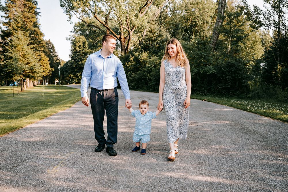 Family of three walking hand-in-hand on a park path, parents holding toddler boy between them on sunny day.