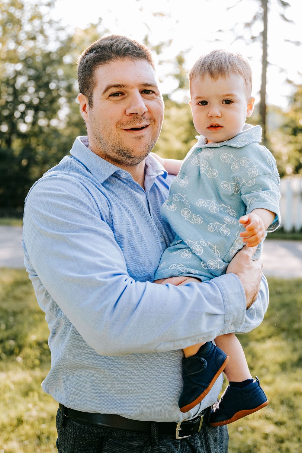 A smiling man in a blue shirt holds a baby boy dressed in light blue outdoors in a sunny park.
