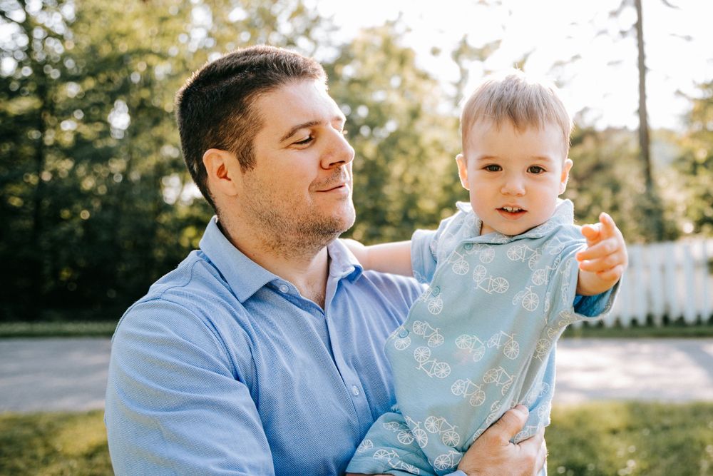 A man in a blue shirt holds a smiling baby outdoors near trees and a white fence.