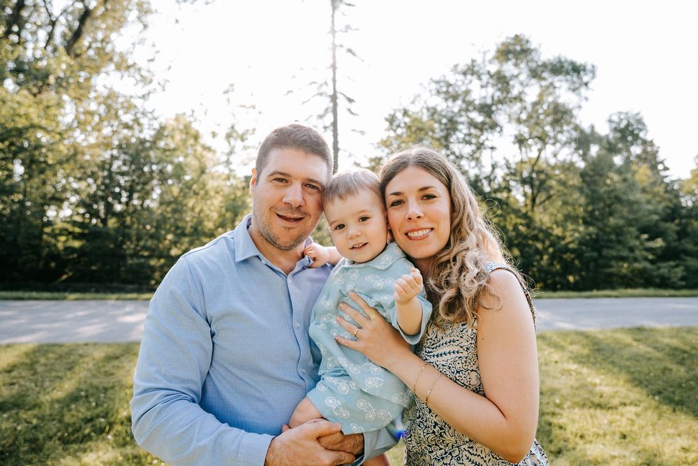 Happy young family with baby posing outdoors in sunny park setting with green trees in background.