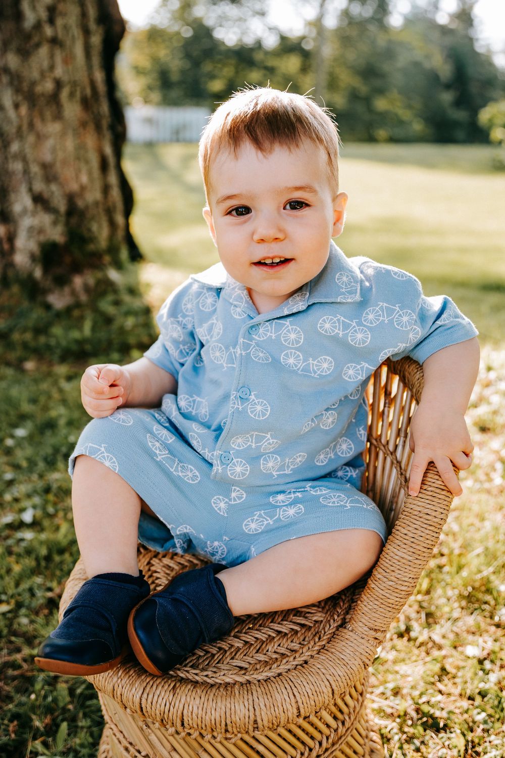 Adorable baby boy in blue bicycle print romper sitting in wicker chair outdoors in sunny green park.