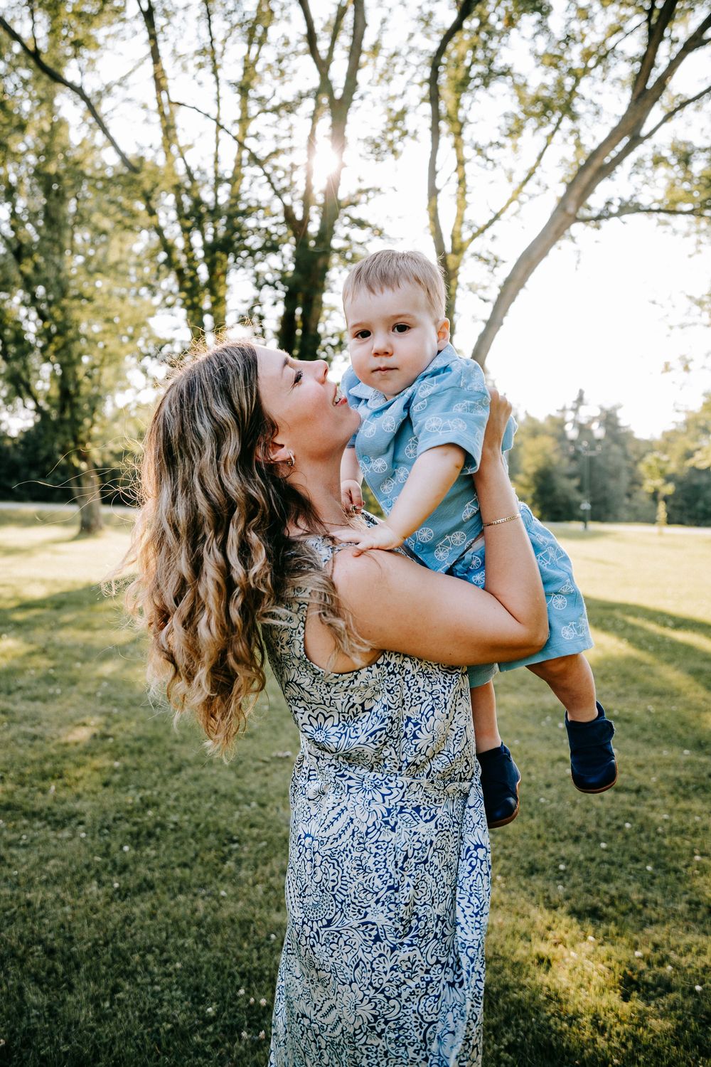 A mother lovingly kisses her toddler boy while lifting him up outdoors in a sunny green park.