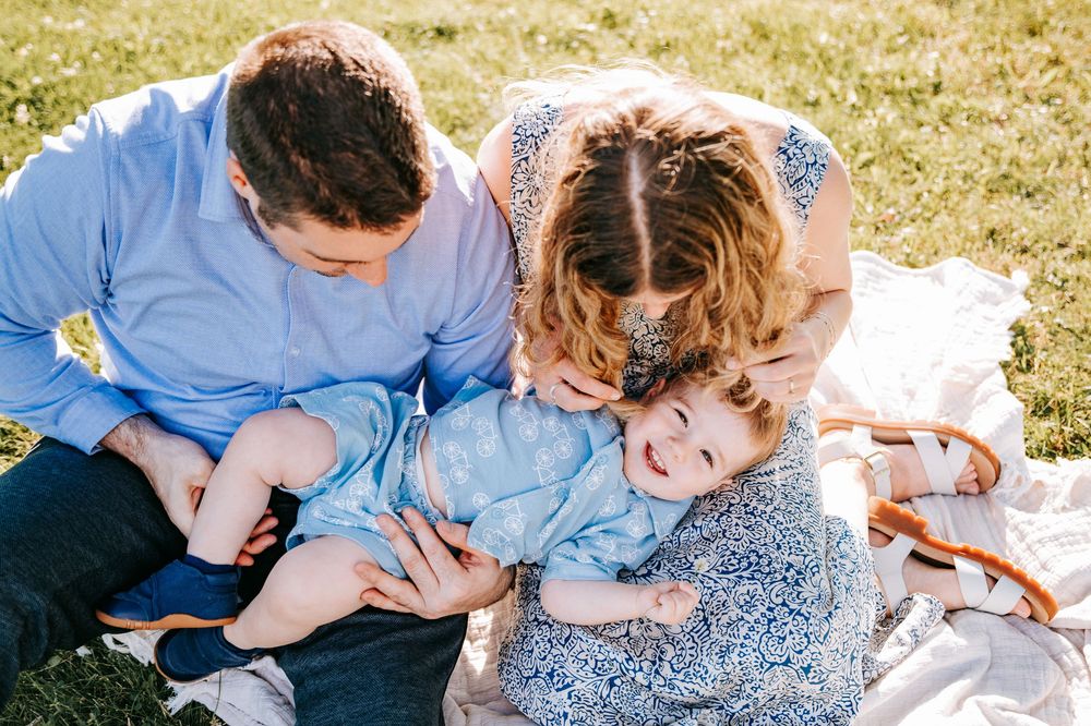 A happy family of three enjoys a sunny outdoor picnic, with parents smiling at their laughing baby on a blanket.