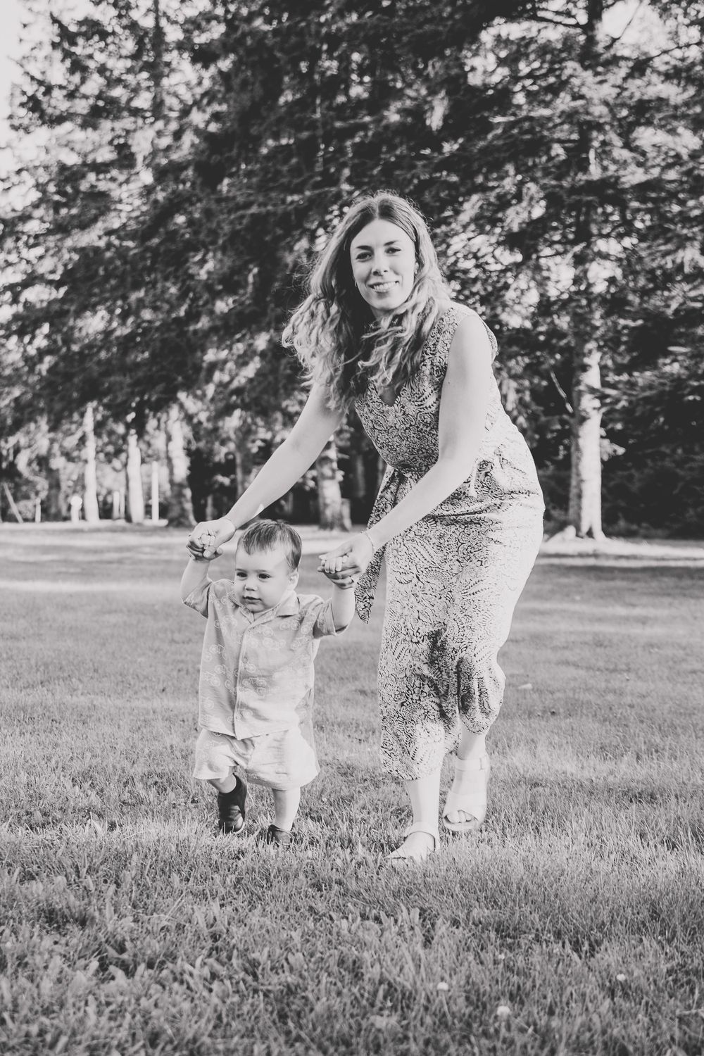 Black and white photo of a mother helping her toddler take first steps on green grass in a sunny park.