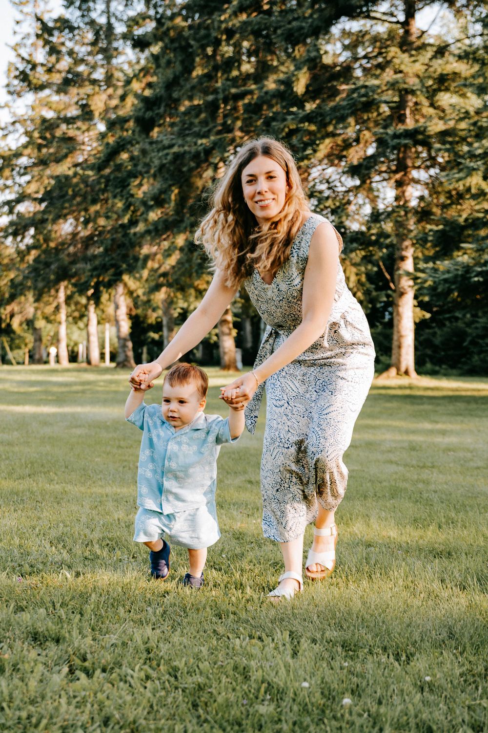 A mother holds her toddler's hands while walking on green grass in a sunny park with tall trees in the background.