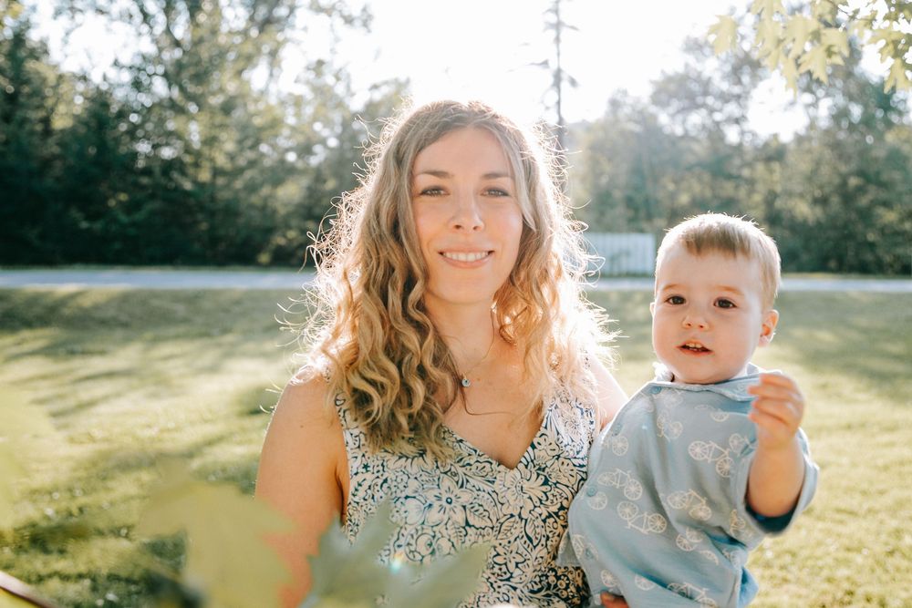 A smiling woman with curly blonde hair holds a baby outdoors in a sunny green park setting.