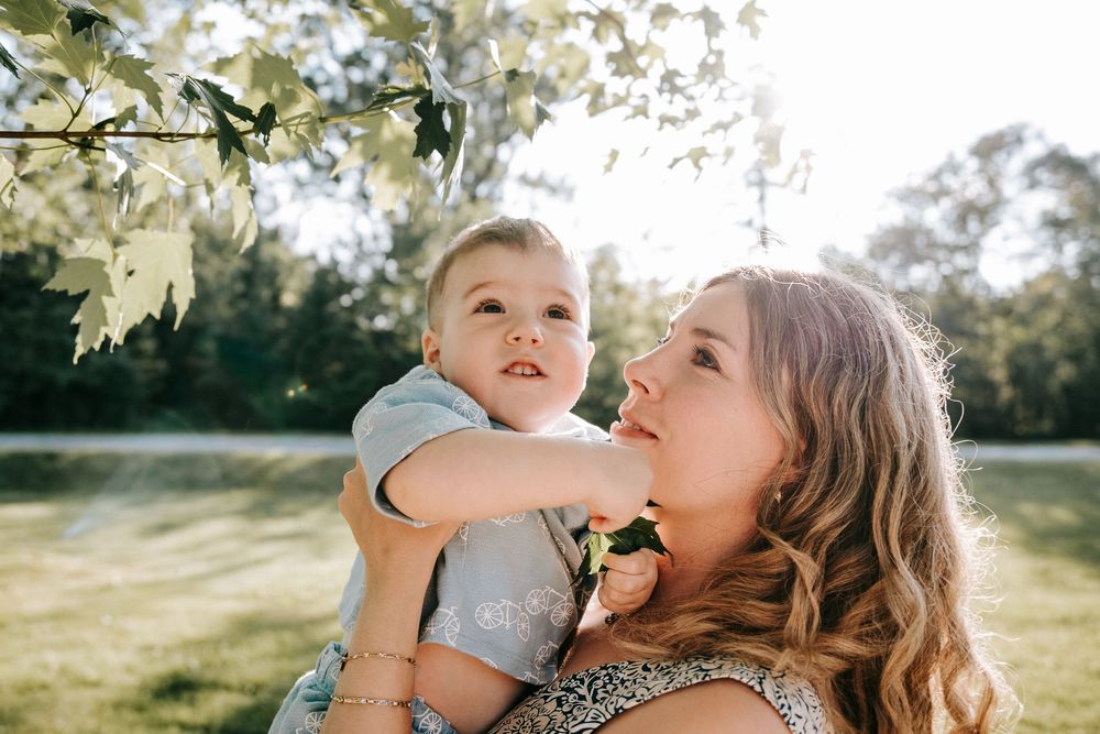 A mother holds her toddler son outdoors under white blossoms in golden sunlight.