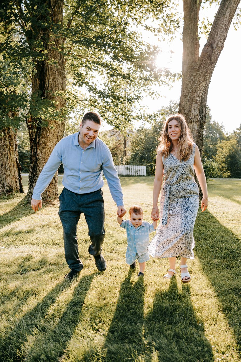 Happy family with toddler holding hands walking in sunny park with large trees and green grass.