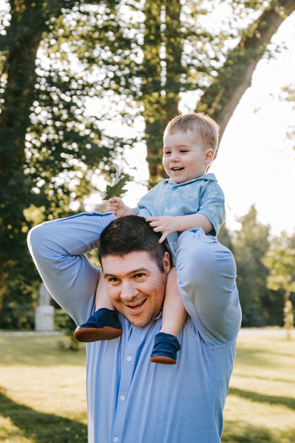 A smiling father carries his laughing toddler son on his shoulders in a sunny green park, both wearing blue shirts.