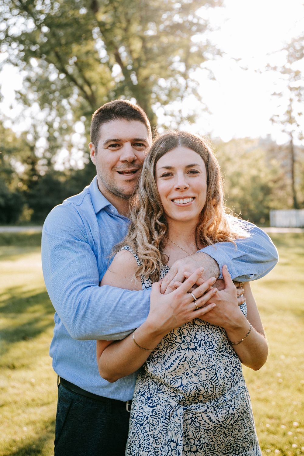 A smiling couple embraces outdoors in a sunny park, man in blue shirt hugging woman in floral dress.