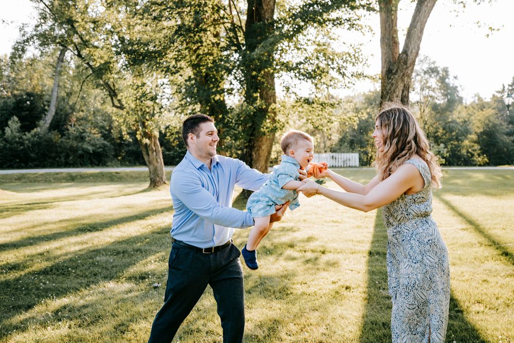 A couple plays with their baby outdoors in a sunny green park, with the man lifting the child toward the woman.