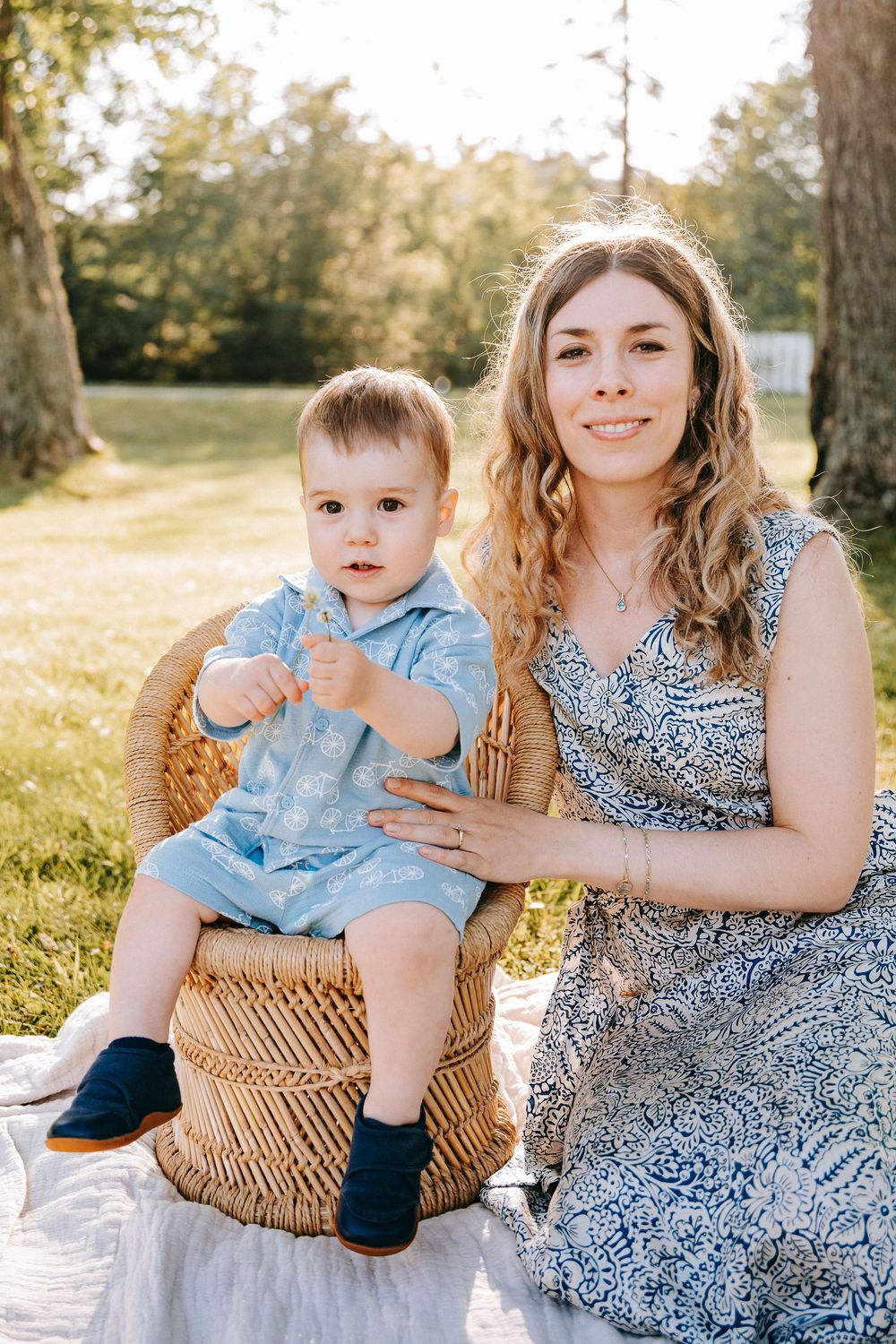 A young mother in a blue dress sits on grass with her toddler boy in a blue outfit on a wicker basket outdoors.