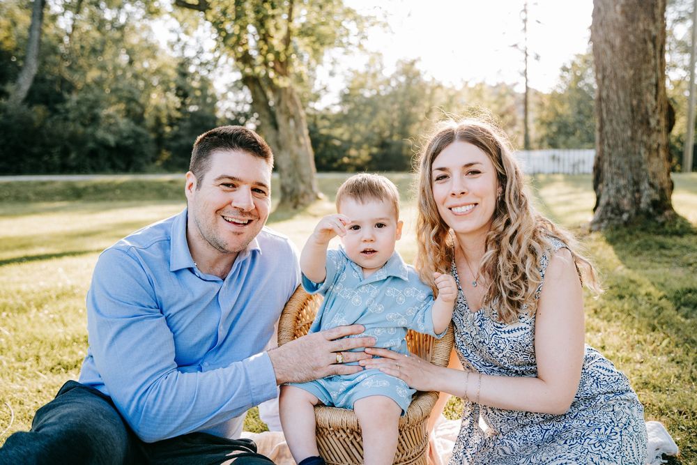 Happy family of three sitting outdoors on grass, parents smiling with baby boy on sunny day.