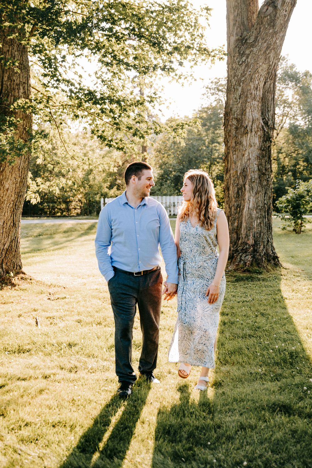 A couple holds hands and smiles at each other while walking on a sunny lawn surrounded by large trees.
