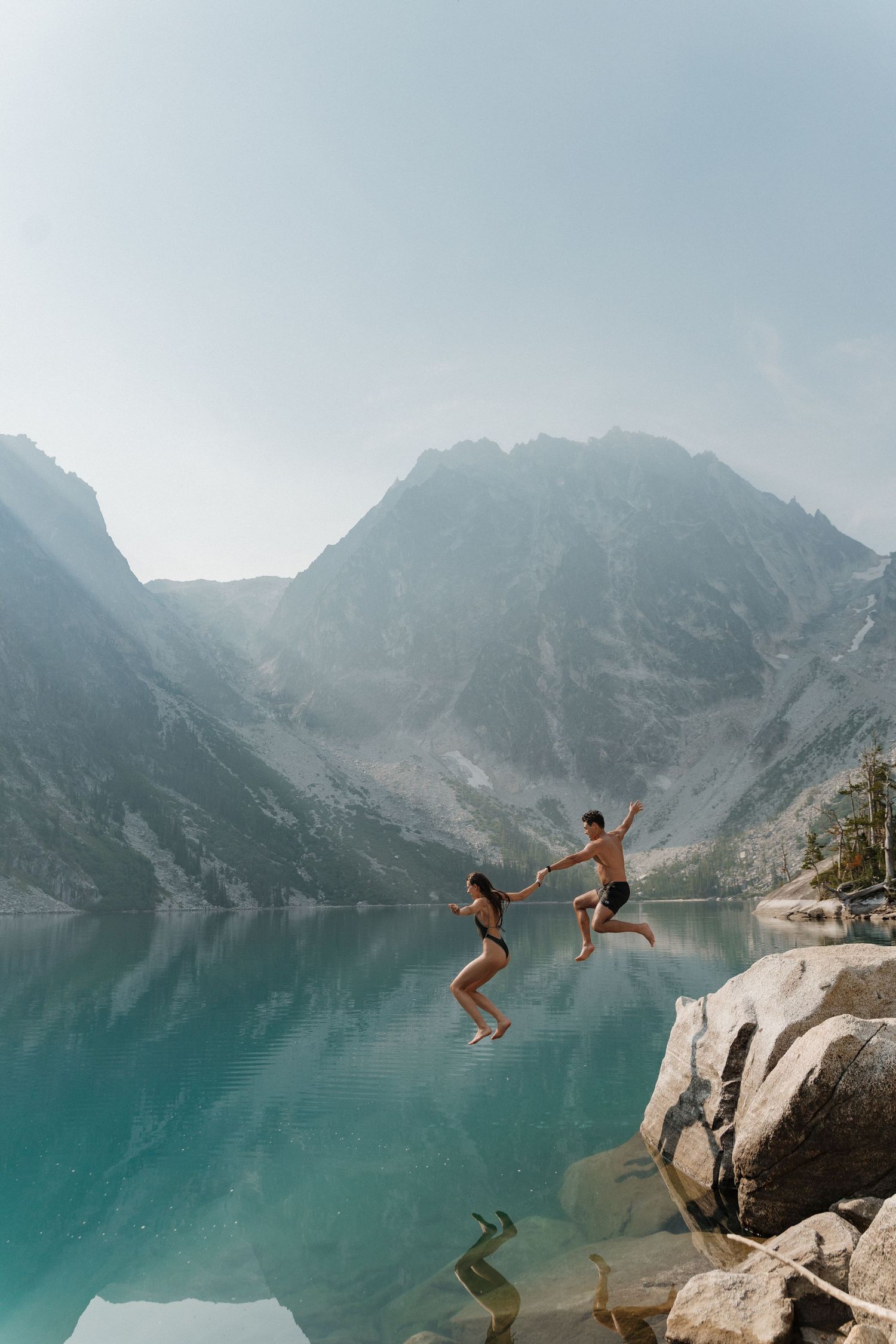 Two people hold hands jumping off a rocky cliff into a turquoise alpine lake surrounded by misty mountains.